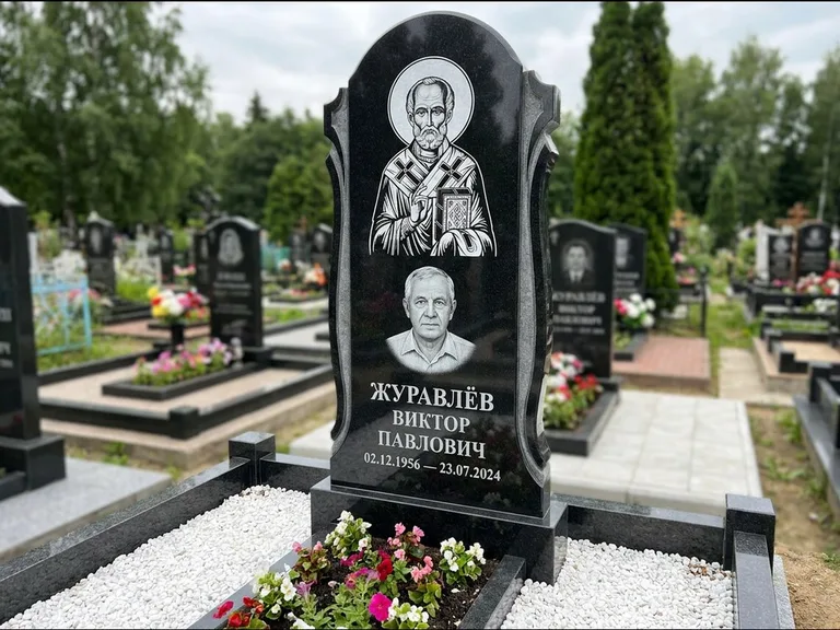 Памятник с гравировкой иконы: стела с образом в полный рост на обороте — гравировка образа Матроны Московской, низкие кусты спиреи в цвету, работа «Лебедева» в Новокузнецке